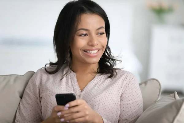 African woman sitting on sofa holds smartphone looking in distance