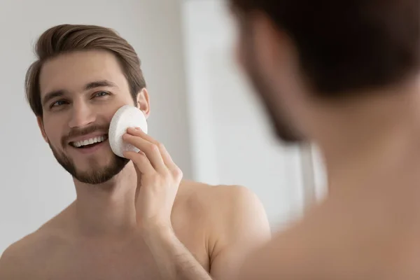 Smiling young man scrub face with sponge in bathroom