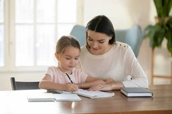 Joven mamá enseñar pequeña hija escritura en cuaderno —  Fotos de Stock