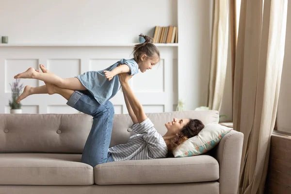 Усміхнена мати тримає маленьку дочку, вдаючи, що летить, лежить на дивані — стокове фото