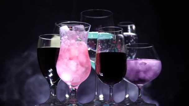Several shaped glasses of various cocktails drinks and ice. Night Club bar table with black gentle fog background and disco lights. Party celebration alcohol concept. Rotation or spinning. 4k. — Stock Video