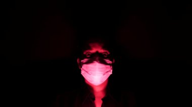 Karanlıkta ve kırmızı ışıkta cerrahi maske takan kadın. Coronavirus 'tan ya da covid 19' dan kişisel karantina izolasyonu. Küresel salgının kıyametleri ve depresyon havası.