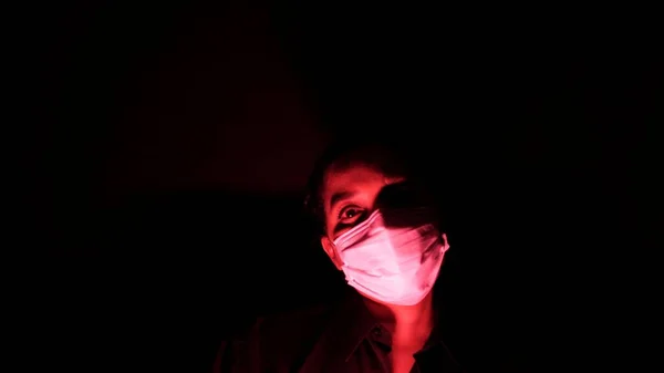 Woman in the surgical medical mask in the darkness and red light. Personal forced home quarantine isolation from Coronavirus or covid 19 concept. Apocalypses and depression mood of global epidemic.