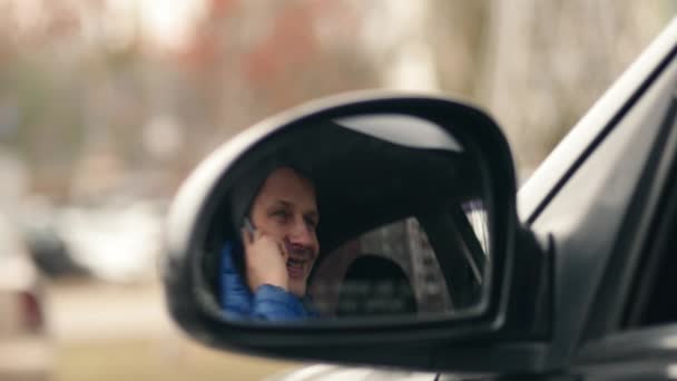 Young man sitting in a car and talking on the phone, shot through the mirror car — Stock Video