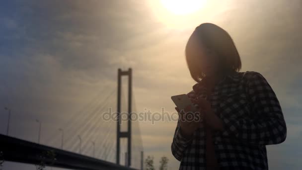 Silhuetten av en flicka i solnedgången med en mobiltelefon-utomhus — Stockvideo