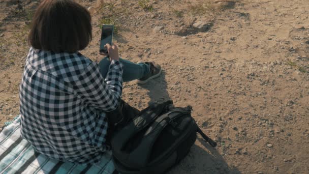 Uma mulher senta-se na areia e lê ao telefone — Vídeo de Stock