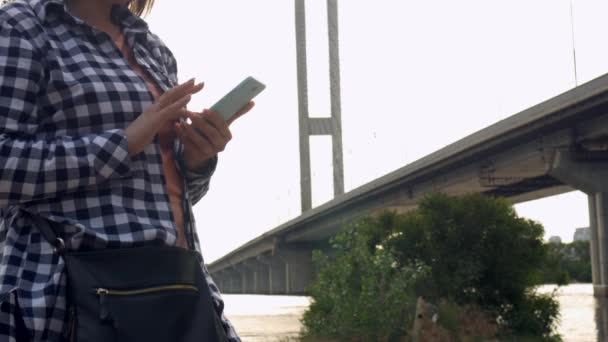 Дівчина на природі отримує SMS на мобільному телефоні на фоні мосту — стокове відео