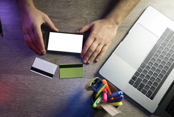 Mãos masculinas segurando smartphone com tela branca, dois cartões de crédito e usando laptop. Conceito de compras online. Vista superior — Fotografia de Stock