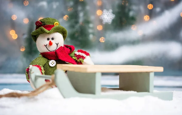 Speelgoedsneeuwpop met sleeën en feestelijke verlichte kerstachtergrond — Stockfoto