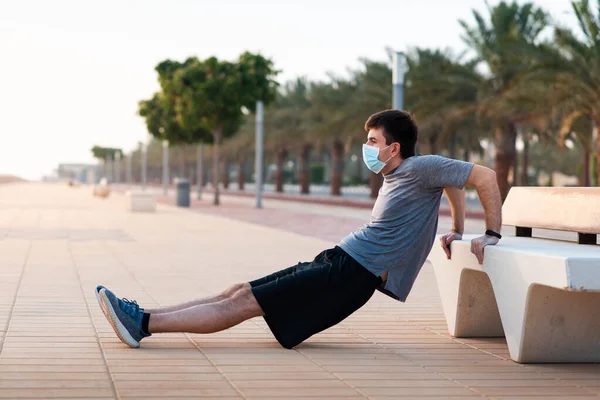 Man doing triceps dips on a bench outdoors and wearing protective surgical face mask