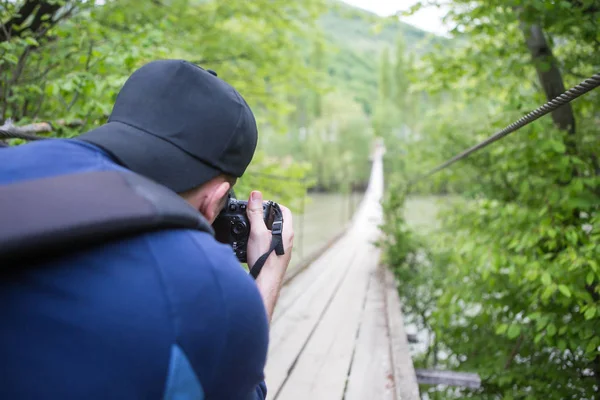 Cestovatel s batohem s fotografii visutý most přes horské řeky s profesionální kamerou — Stock fotografie