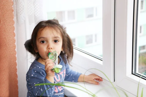 Девочка стоит у окна и ест детское питание — стоковое фото