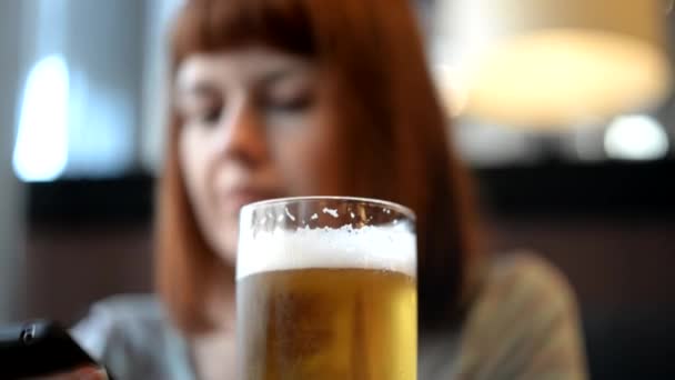 Una chica está sentada en un café bebiendo cerveza y mirando el teléfono. — Vídeos de Stock