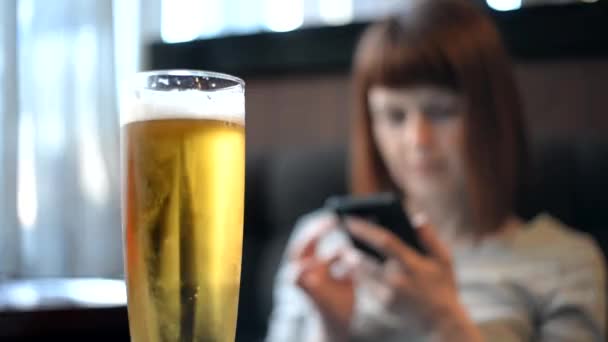 Девушка сидит в кафе, пьет пиво и смотрит в телефон. — стоковое видео