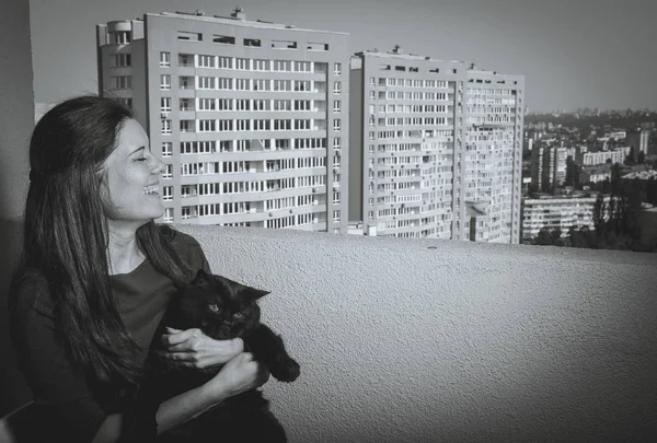 Uma menina com um gato preto olha da varanda para um novo edifício — Fotografia de Stock