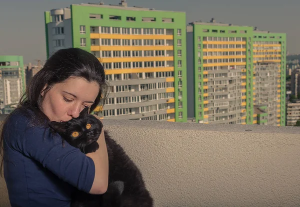 Uma menina com um gato preto olha da varanda para um novo edifício — Fotografia de Stock