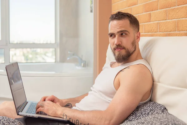 A guy with a beard lies in bed and works behind a laptop — Stock Photo, Image