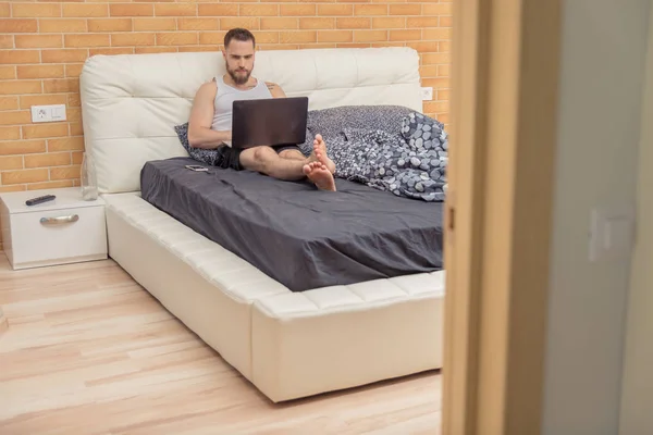 Un tipo con barba yace en la cama y trabaja detrás de un portátil — Foto de Stock