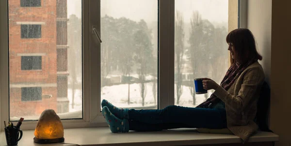 Winter. A girl sits on a window sill and drinks her — Stock Photo, Image