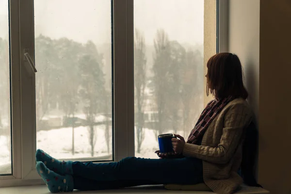 El invierno. Una chica se sienta en un alféizar de la ventana y se la bebe — Foto de Stock