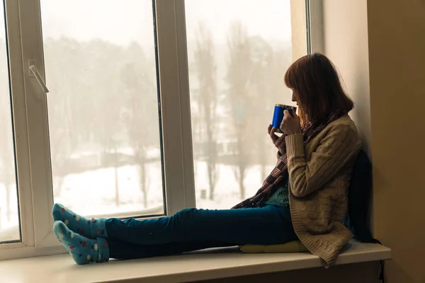 Kış. Bir kız bir pencere pervazına üzerinde oturur ve onu içer — Stok fotoğraf