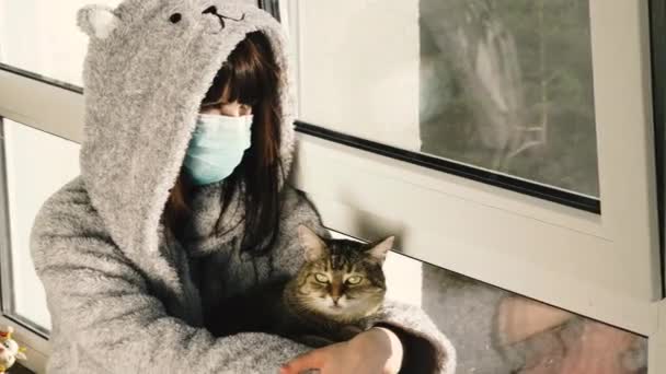 隔離だ 保護医療用マスクの女性が猫と窓辺に座っている — ストック動画
