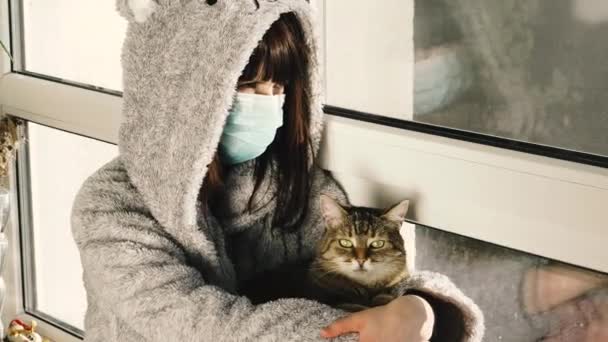 隔離だ 保護医療用マスクの女性が猫と窓辺に座っている — ストック動画