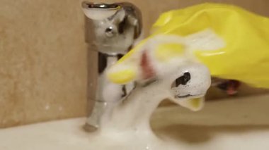 Temizlik. Koruyucu eldivendeki bir el banyodaki musluğu bir bez ve deterjanla yıkar..