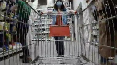 Alışveriş arabası. Koruyucu maskeli bir kadın süpermarkette dolaşıyor. Zaman Bükümüyle Çekildi.