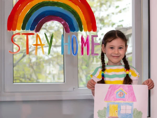 Quarantine fun.Beautiful girl shows a painted house on a background of a window with a painted rainbow.Flash mob society community on self-isolation quarantine pandemic coronavirus. Stay home!let\'s all be well.