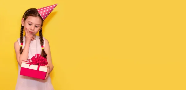 Thoughtful girl in dress looking at gift on hand and thinking about it isolated over yellow.Holiday concept.Copy space for text. Banner