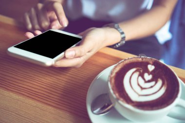 Genç kız beyaz telefon tutarak kafede, boş bir ekran var ve kahve masada hayatı daha fazla yapmak için rahatlatıcı. Mutlu bir yaşam için teknoloji kullanımı.