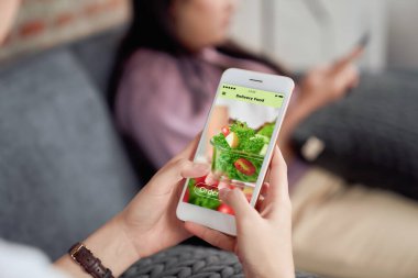 Evdeki kanepede salata siparişi vermek için akıllı telefon kullanan ve uygulama ekranına dokunan bir kadın..