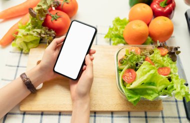 Akıllı telefonlu boş bir ekran, domatesli salata kasesi ve evdeki masada yeşil yapraklı sebzeler tutan bir kadın.. 