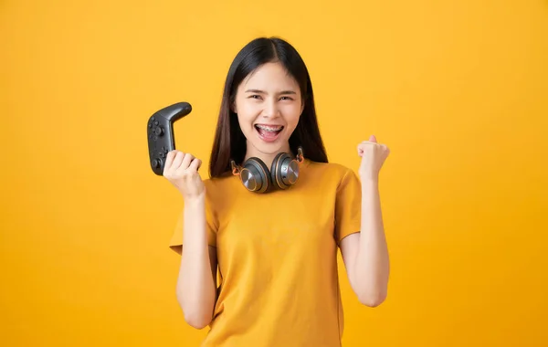 Cheerful beautiful Asian woman in casual yellow t-shirt and playing video games using joysticks with headphones on orange background.