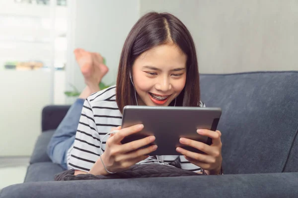 Happy young Asian woman holding a digital tablet and using online social lifestyle on sofa in the home. Technology for communication concept.