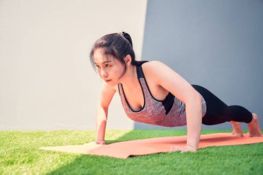 Asyalı kadın yeşil çimlerin üzerinde turuncu bir paspasın üzerinde şınav çekiyor. Refah kavramı.