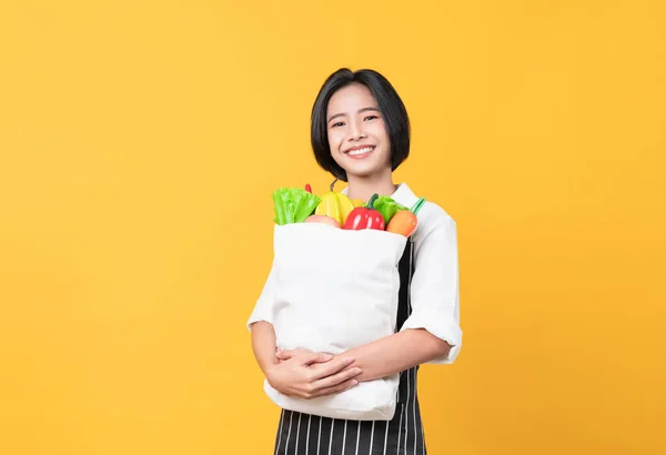エプロン姿のアジア人女性とオレンジを背景に野菜や果物を入れた白いキャンバスバッグを持って立つ — ストック写真