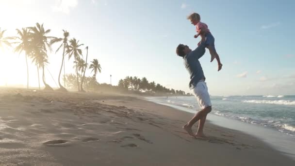 日落时 爸爸把一个孩子扔在大海里 父亲和儿子周末在美丽的热带海滩上玩耍 那里有棕榈树 快乐的童年家庭不完整 — 图库视频影像