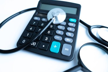 Tıbbi harcamalar ya da blog başlığı ve başlığı için sağlık sigortası kavramı. Stetoskop, beyaz hesap makinesi ve tahta arka planda büyüteç..