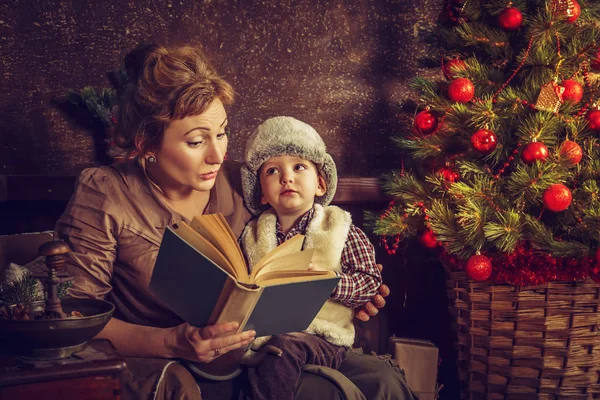 Moeder leest aan zijn zoon het boek in de buurt van een kerstboom. — Stockfoto