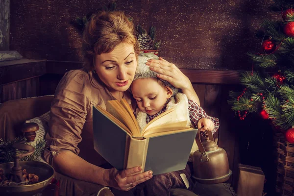 Matka se čte jeho syn kniha poblíž vánoční stromeček. — Stock fotografie