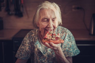 Very elderly woman eating a piece of pizza at home. clipart