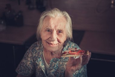 Very elderly woman eating a piece of pizza at home. clipart