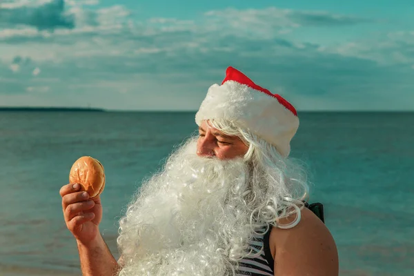 Święty Mikołaj na plaży, jedzenie hamburgera. — Zdjęcie stockowe