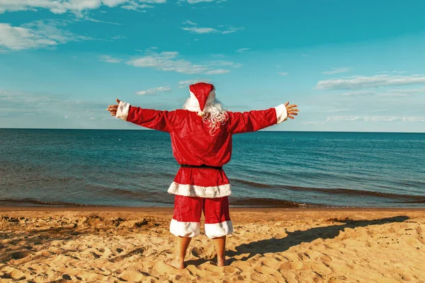 Santa Claus is standing on the beach with arms outstretched.
