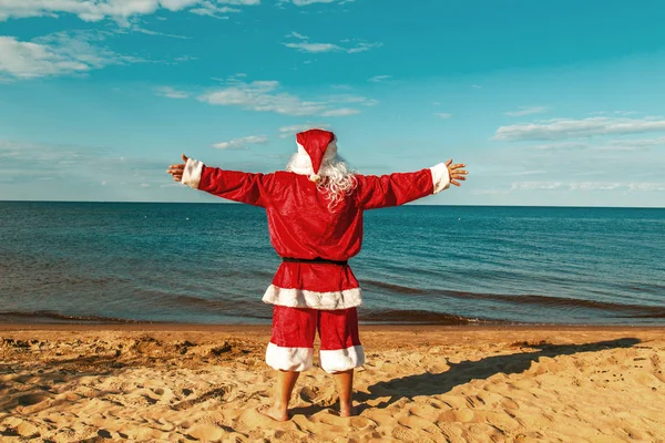 Santa Claus is standing on the beach with arms outstretched. Royalty Free Stock Images