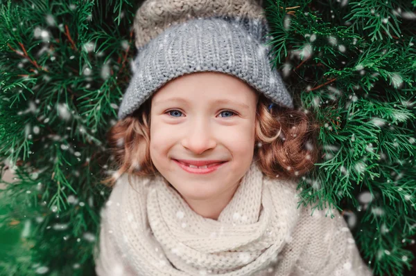 Close up winter portrait of happy child girl wearing warm knitted hat and scarf with Christmas tree on background — Stock Photo, Image