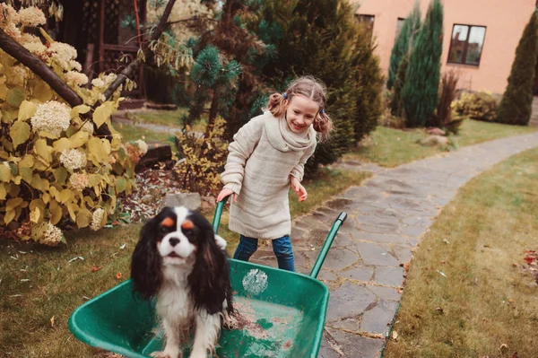 Happy funny child girl riding her dog in wheelbarrow in autumn garden, candid outdoor capture, kids playing with pets — Stock Photo, Image