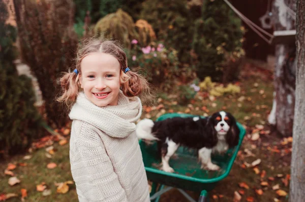 Happy funny child girl riding her dog in wheelbarrow in autumn garden, candid outdoor capture, kids playing with pets — Stock Photo, Image